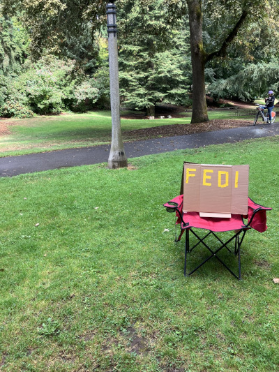 A red folding camping chair sitting on the grass in a park.
             Paved walkway and concrete lamppost behind the chair.
             The chair holds a hand-made cardboard sign that spells out "FEDI" with strips of orange tape.
